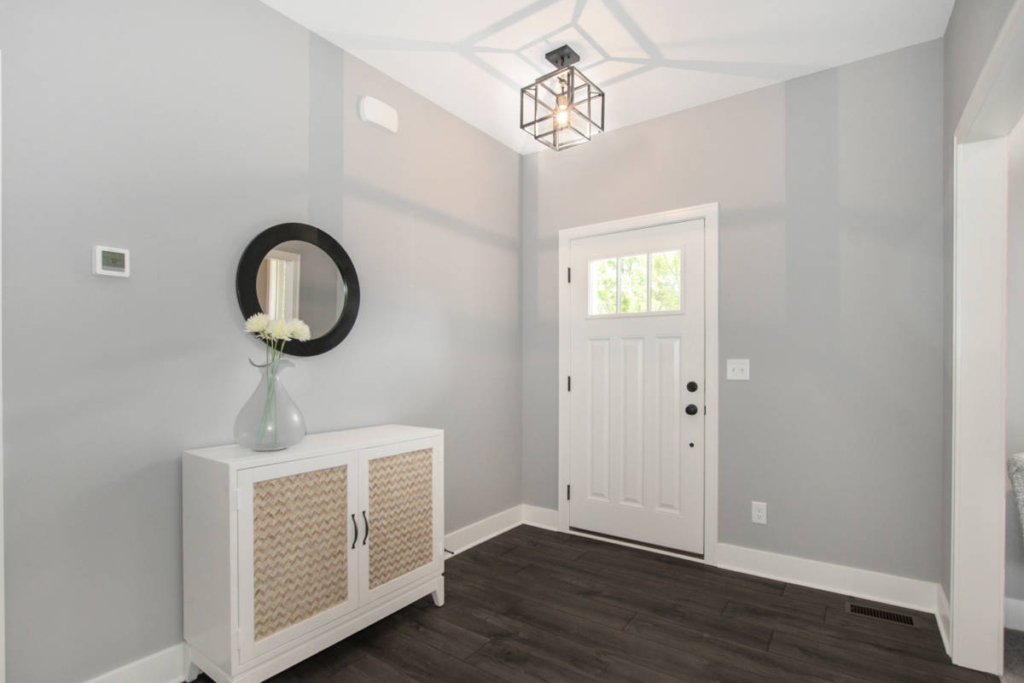 A minimalist entryway with a white door, a small cabinet, a circular mirror, and a vase with white flowers on the cabinet. The floor is dark wood, and the walls are light grey.