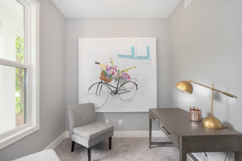 A gray room with a gray upholstered chair, a dark gray desk with a gold lamp and colored pencils, a window, and a large wall painting of a bicycle with flowers.