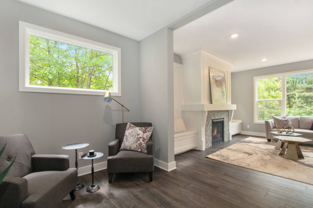 A modern living room with gray walls, dark hardwood floors, and large windows. Two gray armchairs with a side table are on the left; a fireplace and seating area are on the right.