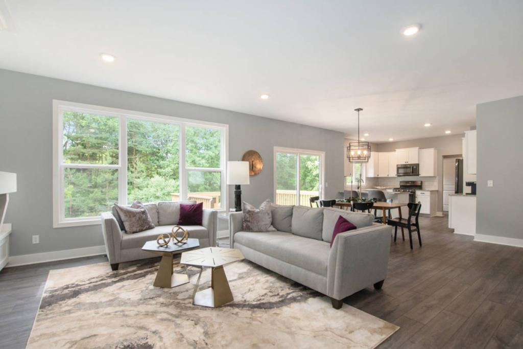 Modern, open-concept living room with two gray sofas, a marble-patterned rug, and a kitchen area in the background. Large windows provide natural light, and contemporary decor is featured throughout.