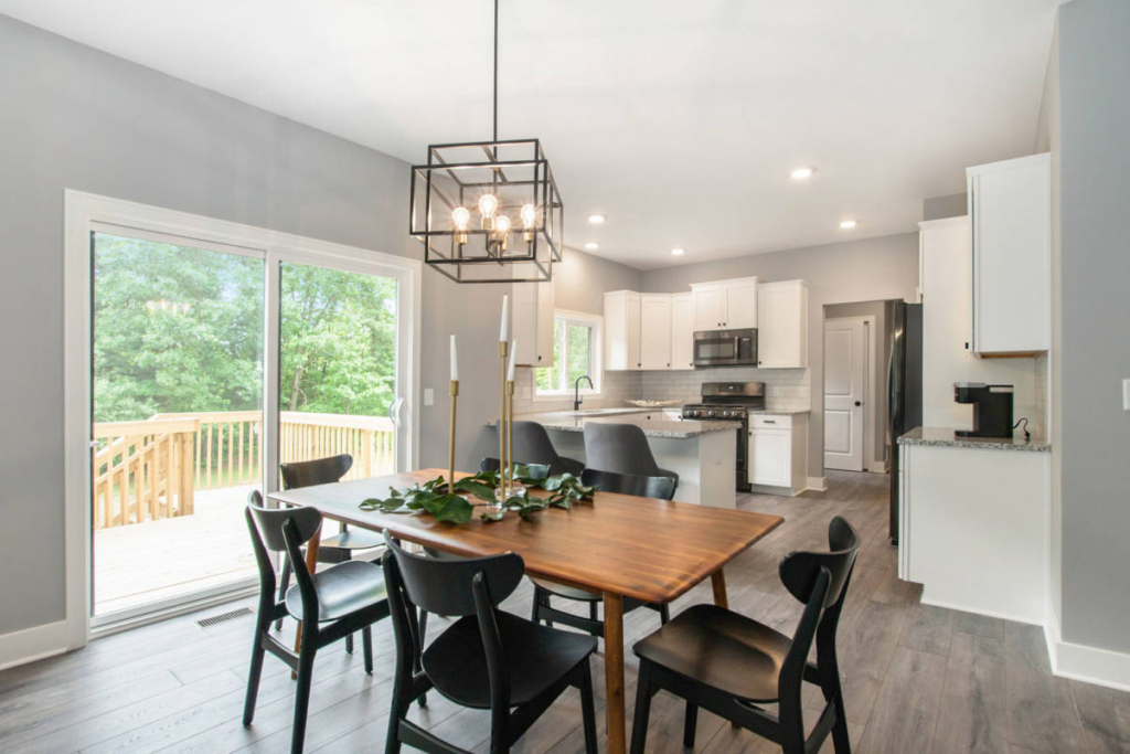 A modern dining area with a wooden table, black chairs, and a decorative centerpiece. The room features a large window overlooking a deck and an adjoining kitchen with white cabinets and stainless steel appliances.