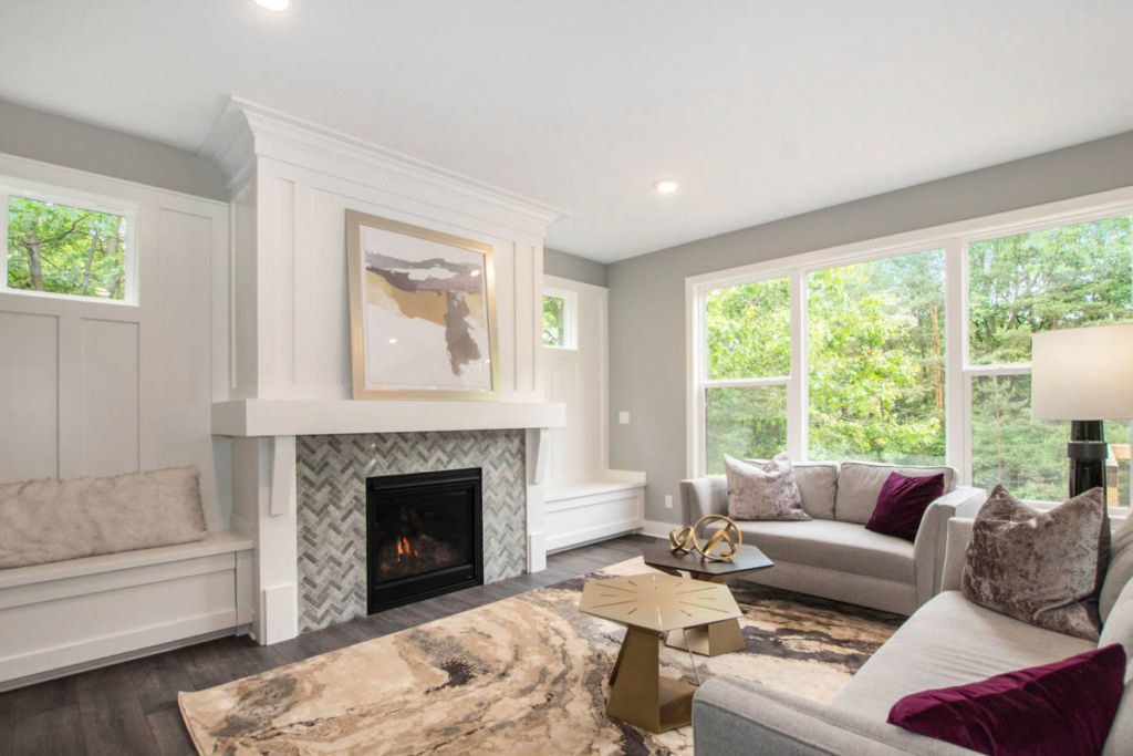 A modern living room with grey sofas, a herringbone-tiled fireplace, large windows, and a geometric coffee table. Natural light brightens the space.