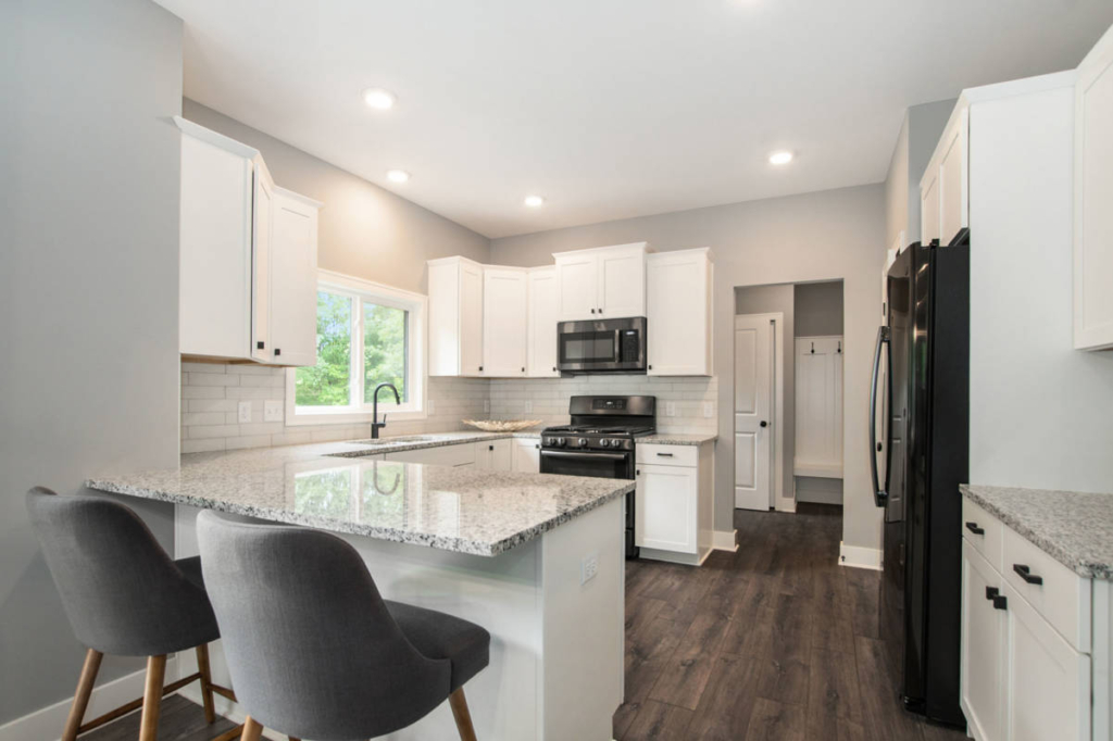 A modern kitchen with white cabinets, a granite countertop island with two gray bar stools, a stainless steel stove, black microwave, and refrigerator, and wooden flooring.