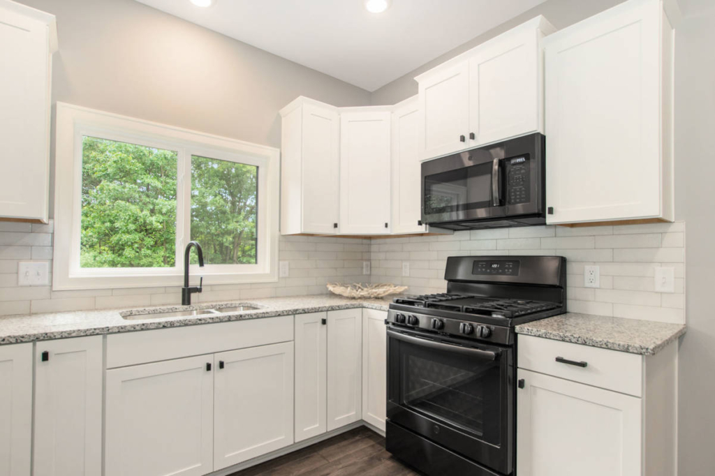 A modern kitchen featuring white cabinets, granite countertops, a black gas stove, and a black microwave. A window with a view of green foliage is above the sink and faucet.