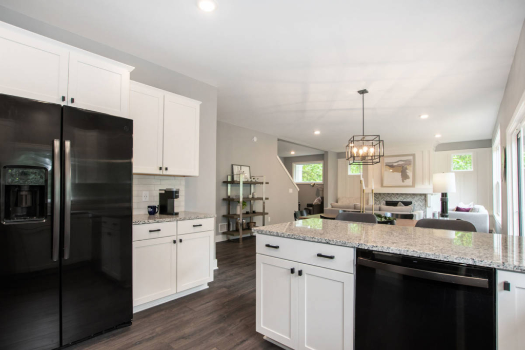 Modern kitchen and living area with white cabinets, black appliances, granite countertops, and hardwood floors. A chandelier hangs above the dining table, and large windows provide ample natural light.