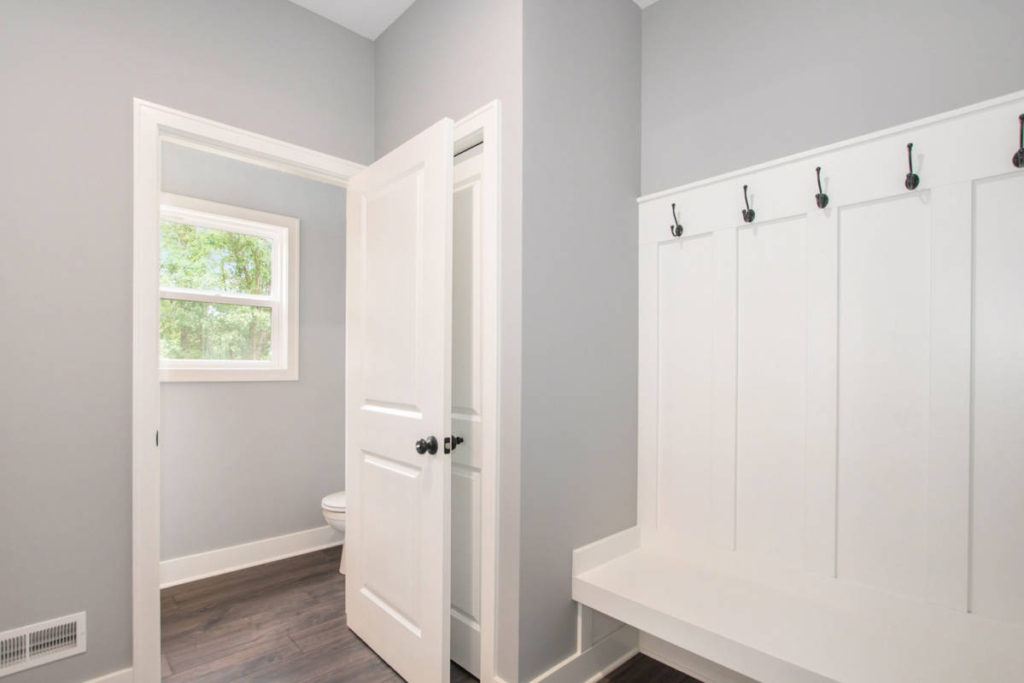 A mudroom with gray walls, a white bench with coat hooks above, dark wood flooring, an open door revealing another room, and a window that shows green trees outside.
