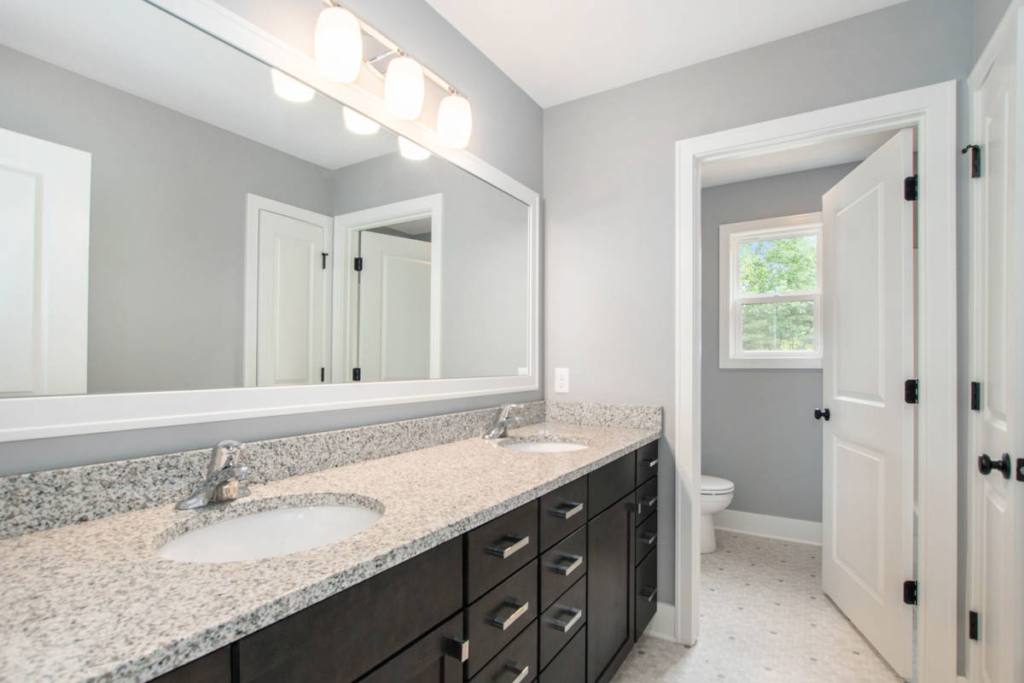 Modern bathroom with a double sink granite countertop, large mirror, and light fixtures. The room features white walls, a window, and a door leading to a toilet area.