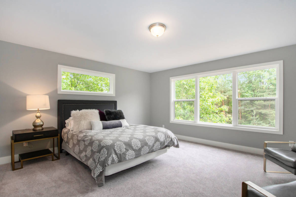 A bedroom with grey walls features a bed adorned with patterned grey and white bedding, a bedside table with a lamp, and large windows overlooking greenery, allowing natural light to fill the room.