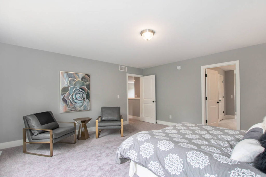 A bedroom with gray walls and carpet, featuring a bed with patterned bedding, two gray chairs, a small table, and a painting on the wall. A doorway leads to an adjacent room.