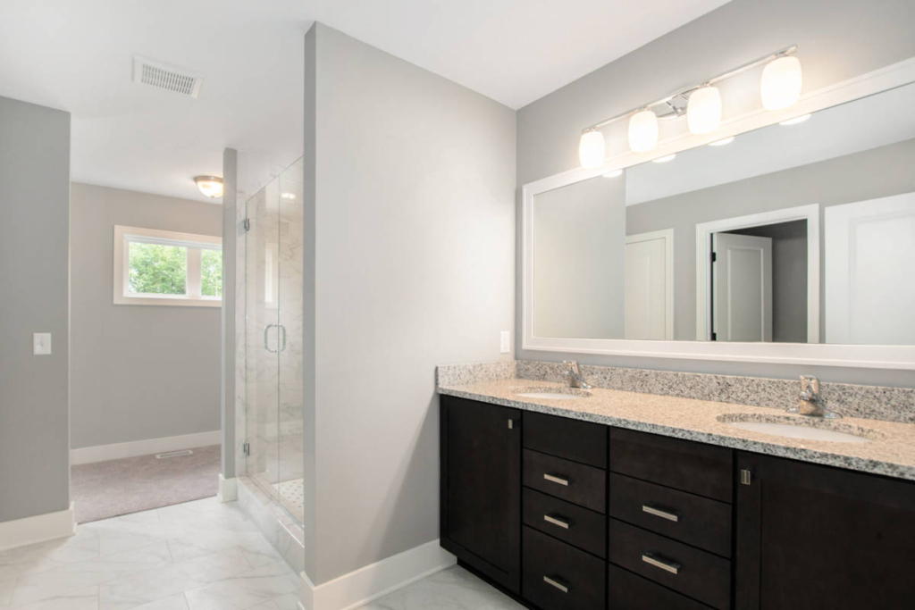 Modern bathroom with a double sink vanity, large mirror, granite countertop, and glass-enclosed shower. Walls are painted light grey and the floor is tiled.