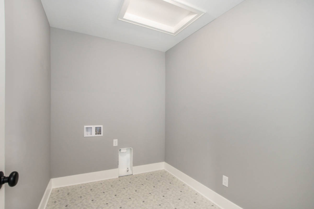 A small, empty laundry room with light gray walls, a linoleum floor, and utility hookups on the left wall beneath a ceiling light.