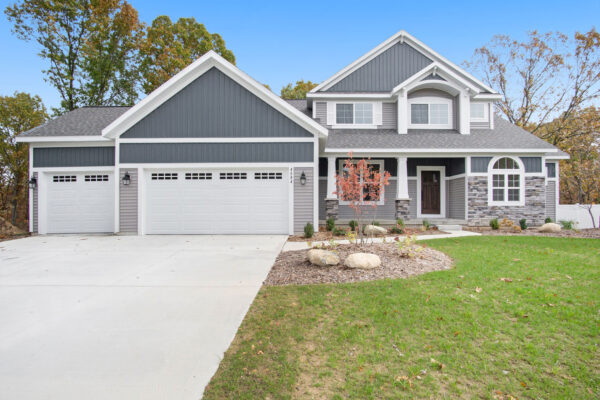 DSC09698 A two-story house with gray siding and stone accents, four-car garage, and a well-maintained lawn. The driveway is paved, and there are trees in the background.