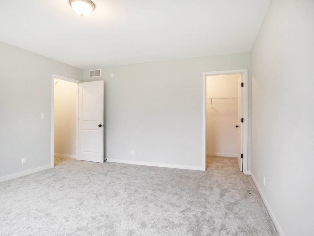 A vacant room with light gray carpet, white walls, two doors, and a small overhead light fixture. One door leads to a walk-in closet, and the other to an adjacent room or hallway.