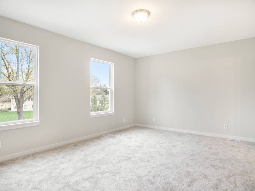 A bright, empty room with beige carpet, neutral colored walls, two windows with outdoor views, and a ceiling light fixture.