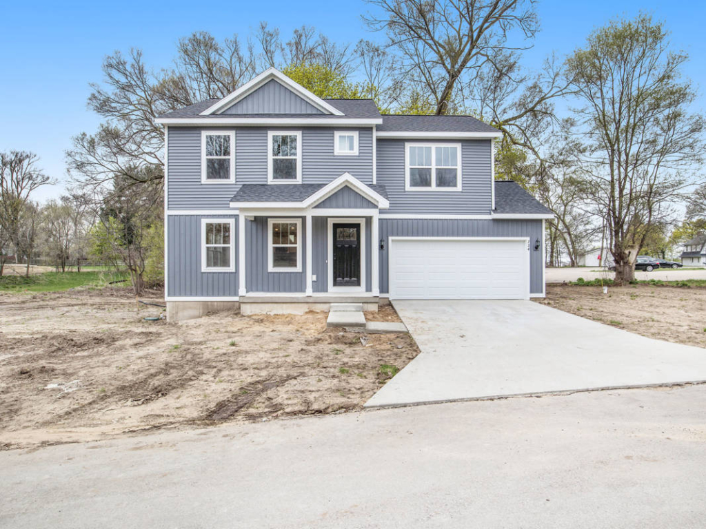 Two-story gray house with white trim, featuring a front porch, large windows, and an attached garage. The property has a concrete driveway and is surrounded by a bare, unfinished yard.