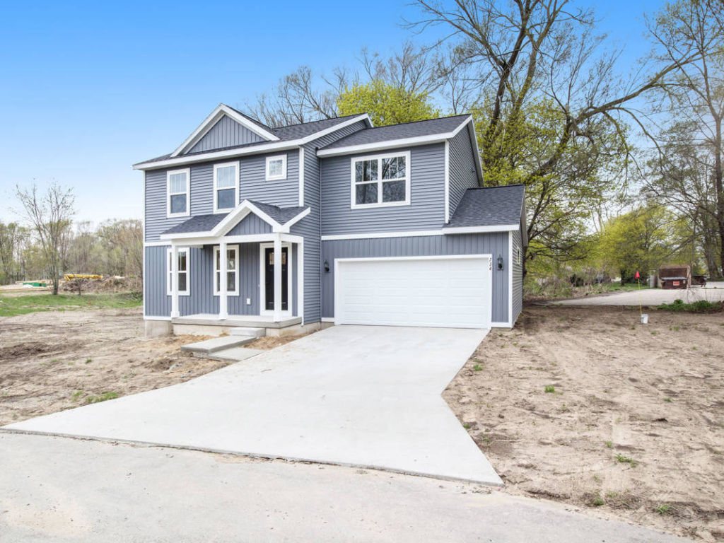 A two-story gray house with white trim, a covered entrance, and an attached garage. The driveway and surrounding yard appear newly constructed and lack landscaping. Trees are present in the background.