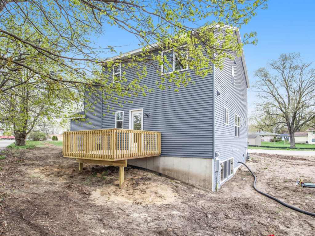 A two-story house with gray siding has a small wooden deck in the backyard, surrounded by unfinished landscaping and trees, under a clear blue sky.
