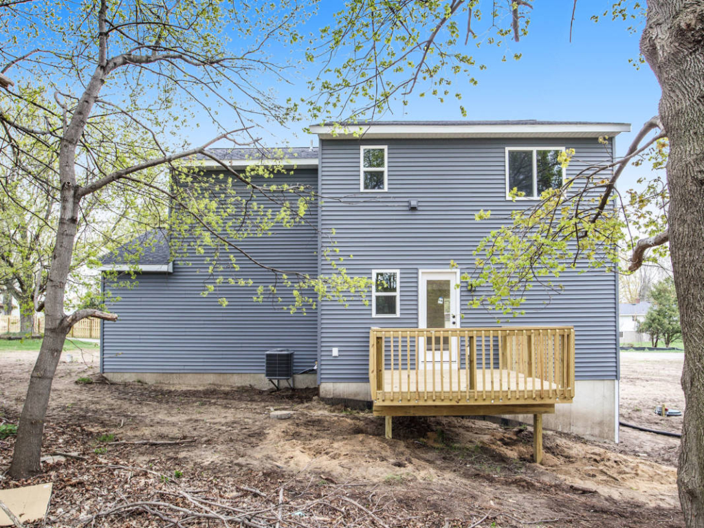 A rear view of a two-story house with gray siding. The yard is bare, with a small wooden deck near the back door and sparse trees nearby. The sky is clear and blue.