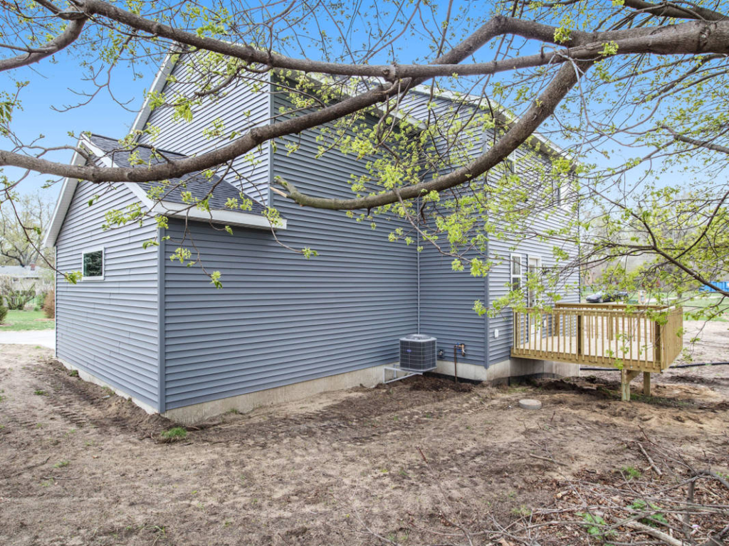 A gray house with side deck and air conditioning unit, surrounded by bare soil and branches with young green leaves in the foreground.