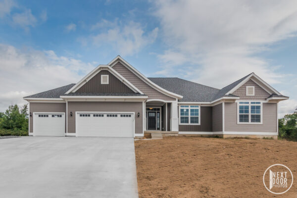 Emerson-IMG_8861 A single-story house with a gray exterior, white trim, and a three-car garage. The front yard is bare soil, and the sky is partly cloudy.