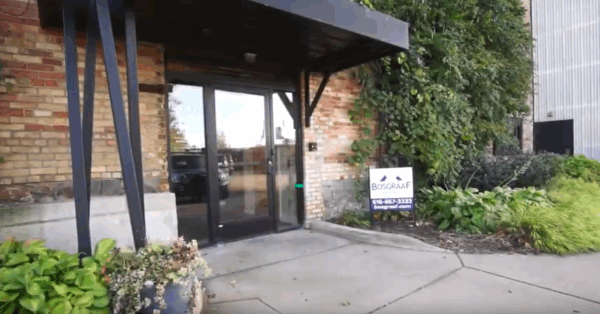 design-center Entrance of a brick building with a glass door, black awning, and surrounding greenery. A sign near the door displays contact information.