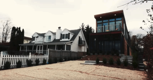 douglas-river-home A modern two-story house with large windows stands beside a traditional white house with a picket fence. Both are set against a cloudy sky.