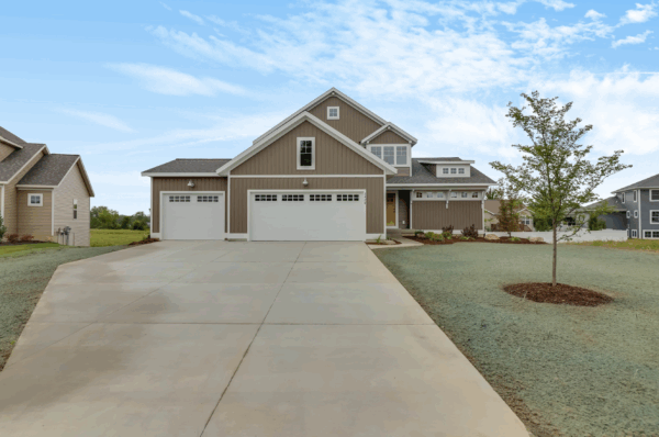 the-duncan Suburban house with a three-car garage, gray siding, and white trim. It has a large driveway, a small tree in the front yard, and a cloudy blue sky overhead.