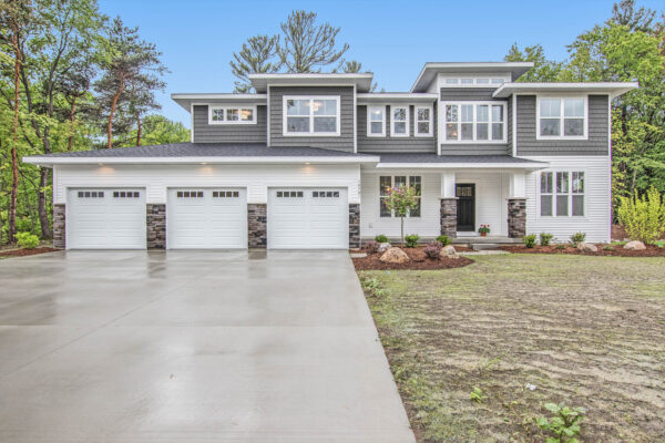 IMG_6044-1 A modern two-story house with a gray exterior, large windows, and a four-car garage, surrounded by trees and a concrete driveway.
