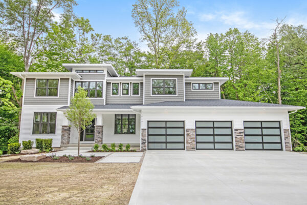 IMG_9992 A modern two-story house with several large windows, a gray and white exterior, a three-car garage, and a driveway. The house is surrounded by trees and has a small front garden.