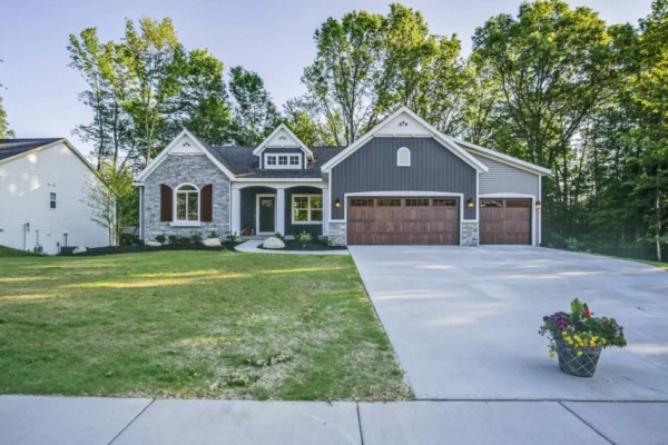 DSC01013Bended A two-story house with grey siding, white trim, and a wooden garage door. It features a large driveway, manicured lawn, and potted flowers in the foreground. Tall trees are visible in the background.
