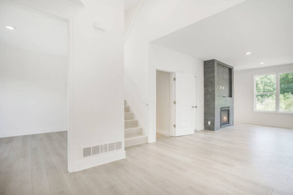 DSC04642 A spacious, unfurnished room with light wooden flooring, a staircase, a white door, a fireplace with a dark accent wall, and a large window letting in natural light.