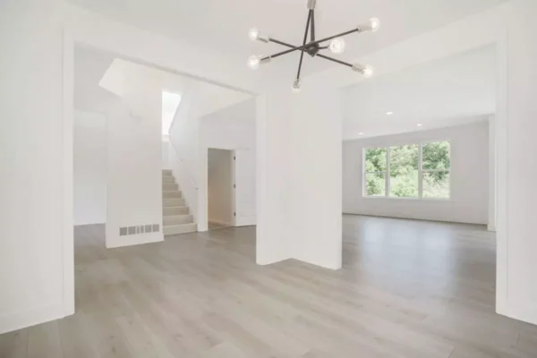 DSC04647 A spacious, bright, and empty modern living room with light wood floors, white walls, large windows, and a contemporary chandelier. There's a view of a staircase and adjacent rooms.