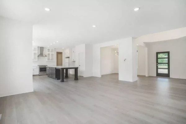 DSC04654 A spacious modern kitchen and living area with light wood flooring, white walls, an island with a black base, stainless steel appliances, recessed lighting, and a door leading to the outside.