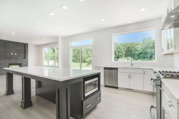 DSC04659 Modern kitchen with a central island featuring a built-in microwave, stainless steel appliances, white cabinets, and large windows allowing natural light. Living room with a fireplace is visible in the background.