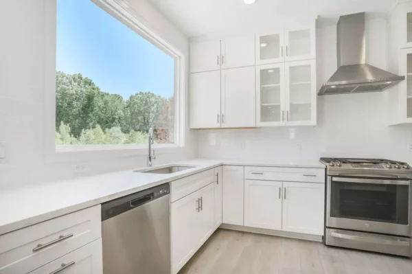 DSC04662 Modern kitchen with white cabinets, stainless steel appliances including a dishwasher and stove, large window with outdoor view, and light wood flooring.