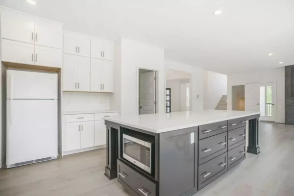DSC04663 Modern kitchen with a large island featuring a built-in microwave, white cabinetry, a refrigerator, and light wood flooring.