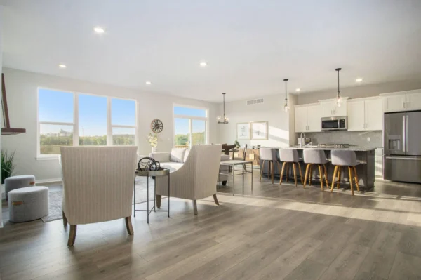 DSC07371 A modern, open-concept living area features a kitchen with an island, bar stools, stainless steel appliances, and a spacious living room with large windows and neutral-toned furniture.