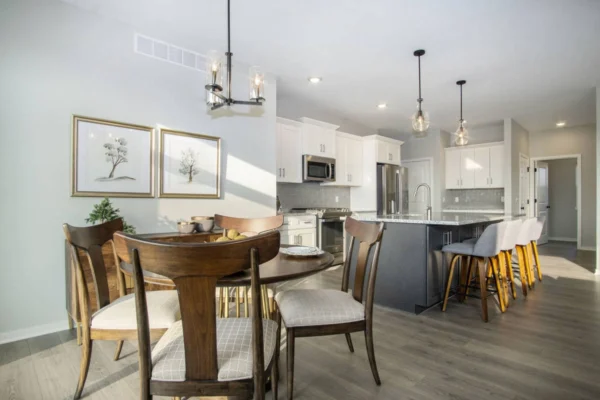 DSC07381 Modern kitchen and dining area with wooden dining table and chairs, white cabinets, stainless steel appliances, and an island with bar stools. Wall art and pendant lights are also visible.