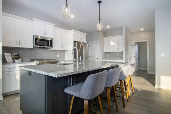 DSC07383 Modern kitchen with gray granite countertops, white cabinets, stainless steel appliances, and a large island with four gray barstools. Hanging pendant lights illuminate the space.