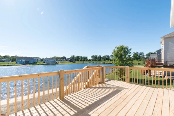 IMG_0267 Wooden deck overlooking a calm lake with houses and trees in the distance under a clear blue sky.