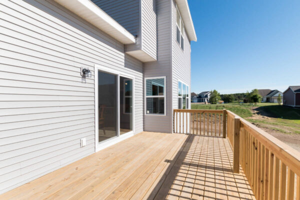 IMG_0268 Wooden deck with railing attached to a gray house under clear blue sky. Houses and lawn visible in the distance.