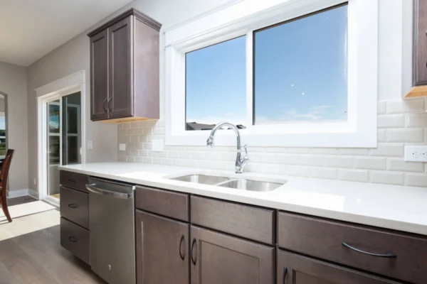 IMG_0273 Modern kitchen with brown cabinets, white countertops, double sink, and large window with a blue sky view.