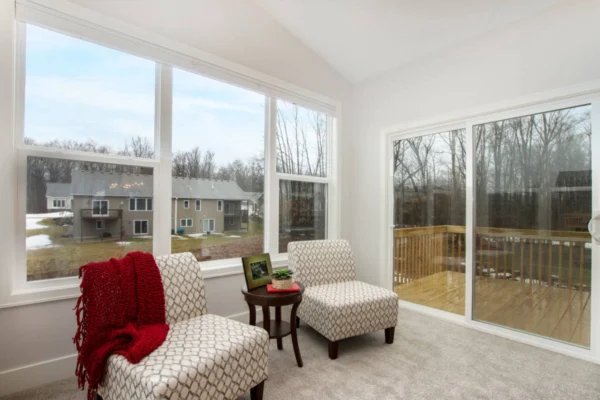 IMG_0293 Bright room with two patterned chairs, a red throw, small table, large windows, and sliding door overlooking a deck.