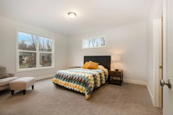 IMG_0299 Bedroom with a bed, geometric-patterned bedding, lamp on a side table, chair, and large windows.