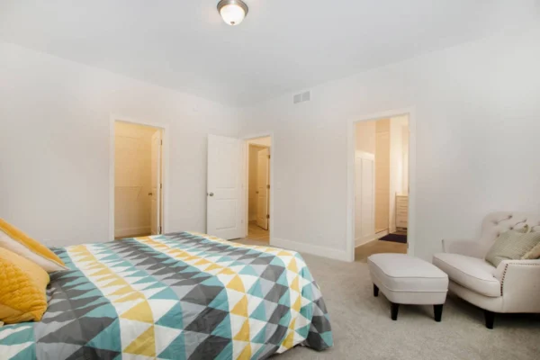 IMG_0305 Bedroom with patterned bedspread, plush armchair, footstool, and open doors to bathroom and closet on a beige carpet.