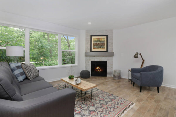 IMG_4678 Living room with a gray sofa, wooden coffee table, armchair, fireplace, floor lamp, and large window providing outdoor view. Rug on the floor and a framed picture above the fireplace.