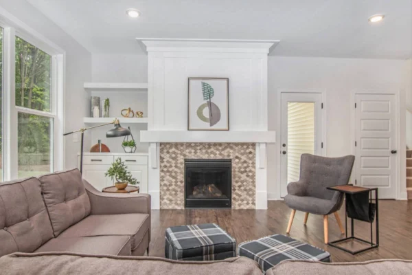IMG_5974 A modern living room featuring a gray couch, patterned ottomans, a fireplace with a tiled surround, and built-in shelves beneath a large window. A framed artwork hangs above the mantel.
