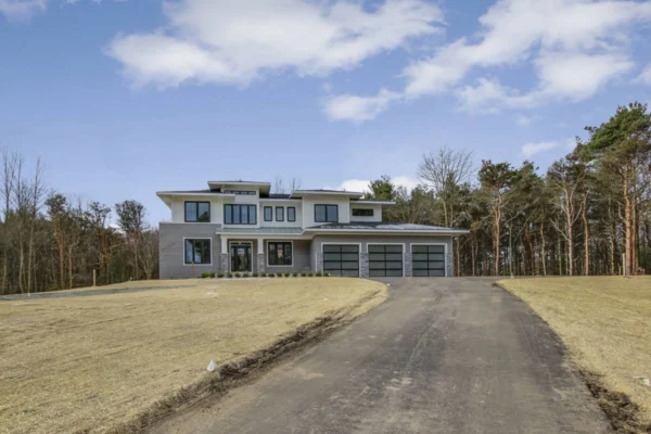 IMG_6378Blended Modern two-story house with large windows, a driveway, and garage, set against a backdrop of trees and partly cloudy sky.