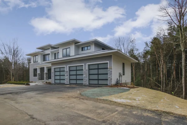 IMG_6381Blended Modern two-story house with large windows and three garage doors, surrounded by trees and a cloudy sky.