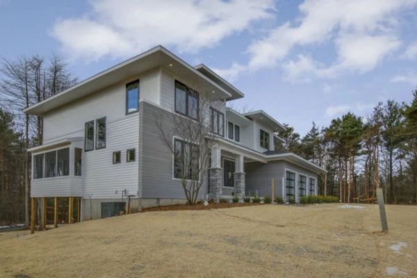 IMG_6384Blended Modern two-story house with gray siding, large windows, and a sloped yard, surrounded by trees under a cloudy sky.